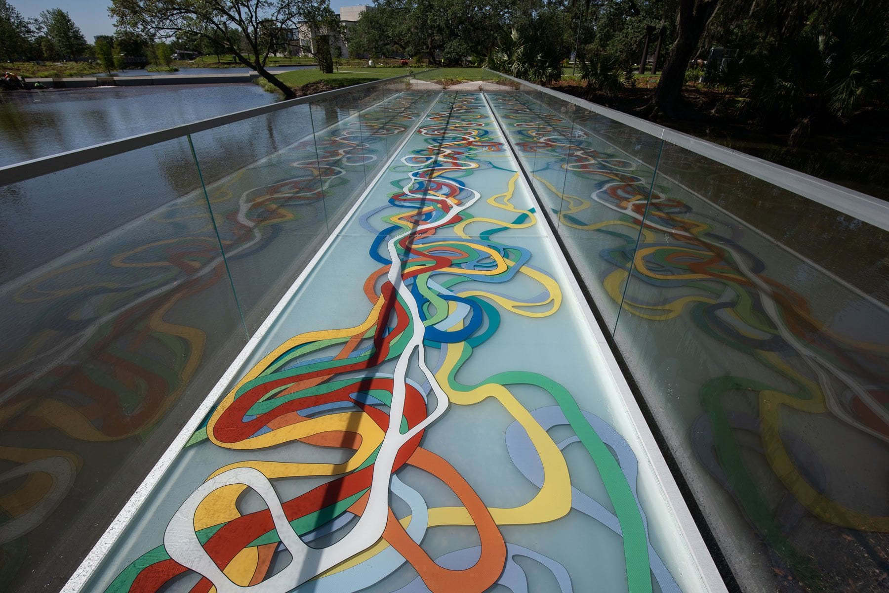 Colorful abstract mural on a glass bridge over water with trees and greenery in the background on a sunny day.