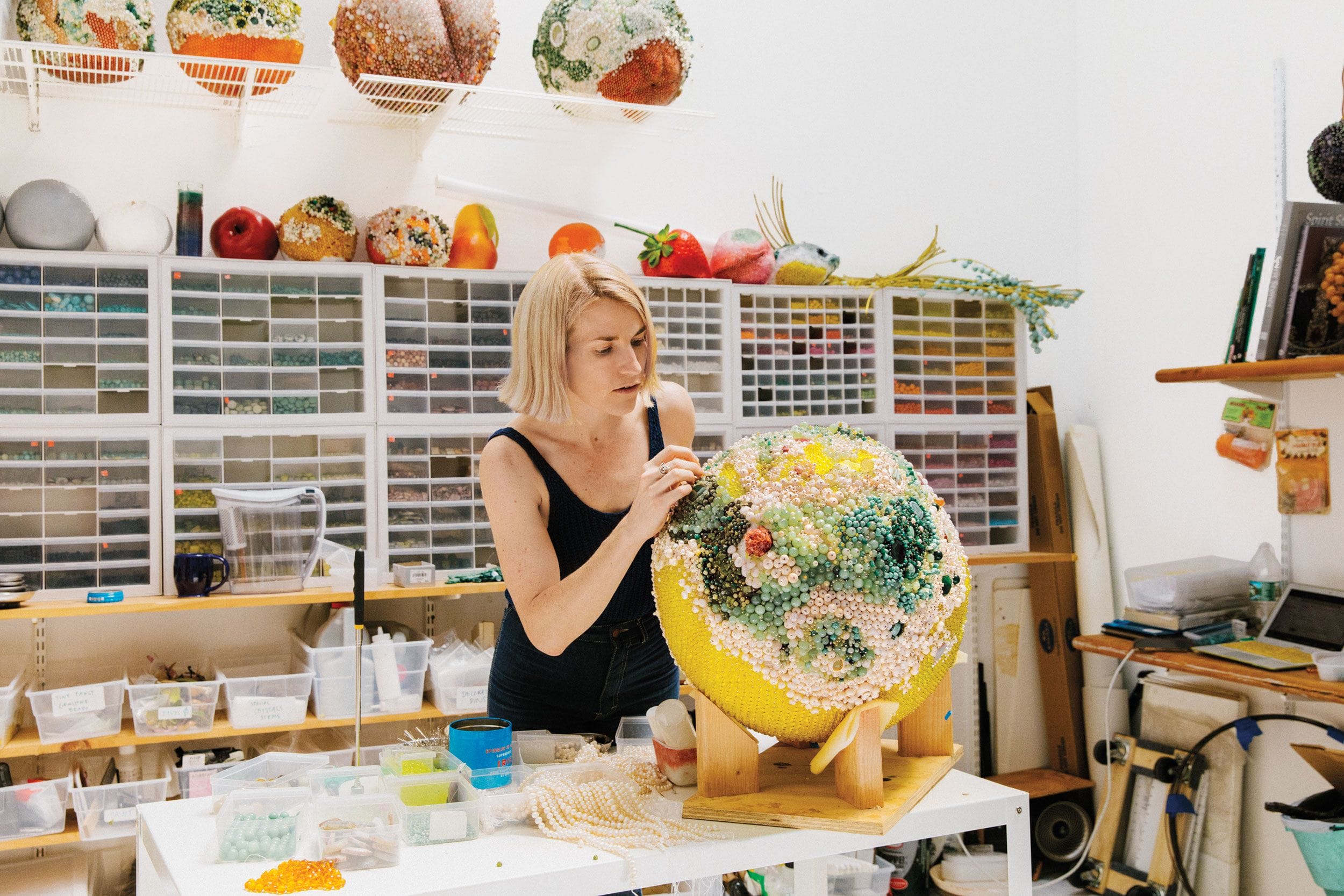 Artist working on a colorful globe sculpture in a studio filled with art supplies and decorated spheres.