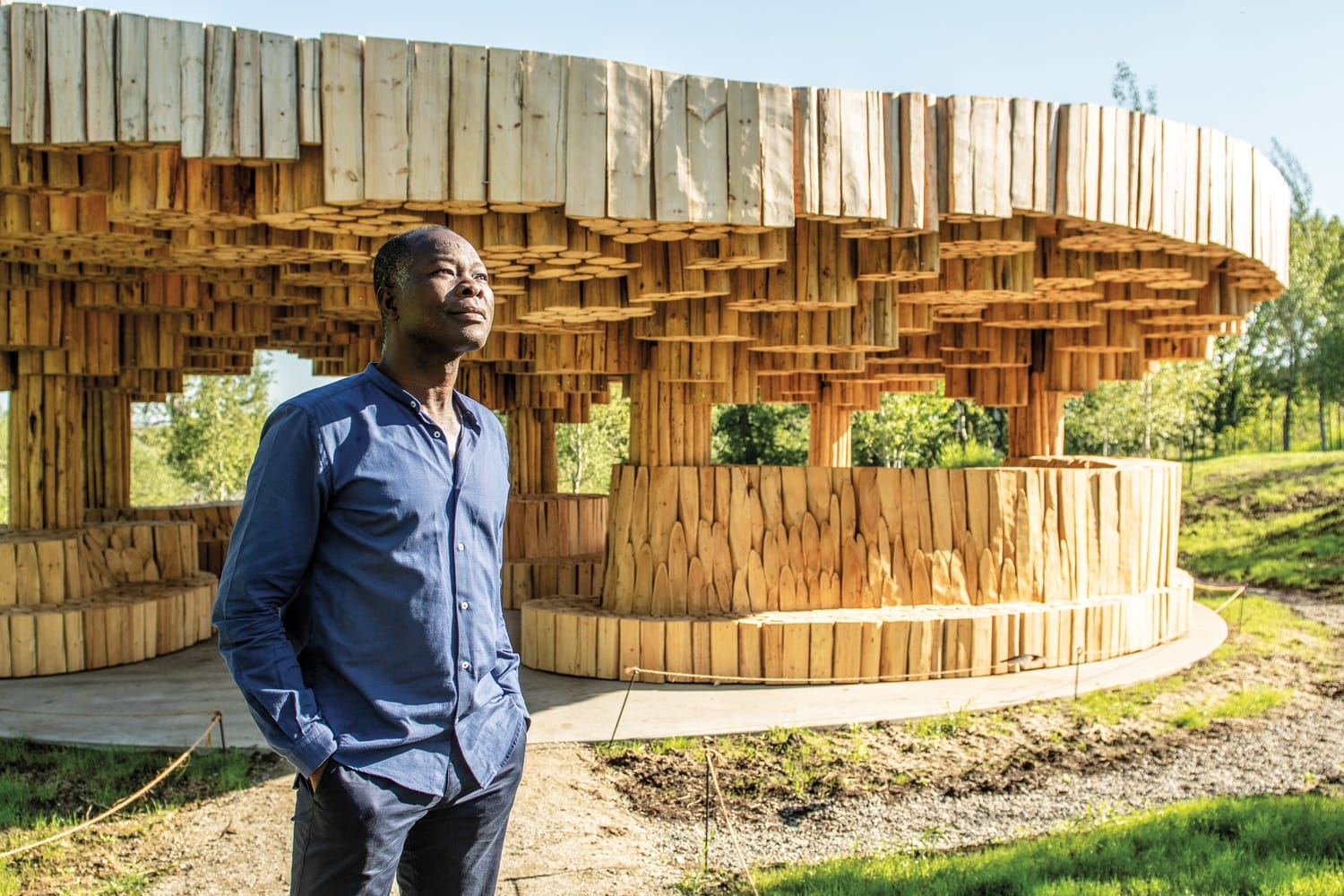Man standing in front of a wooden architectural structure in a grassy outdoor setting.