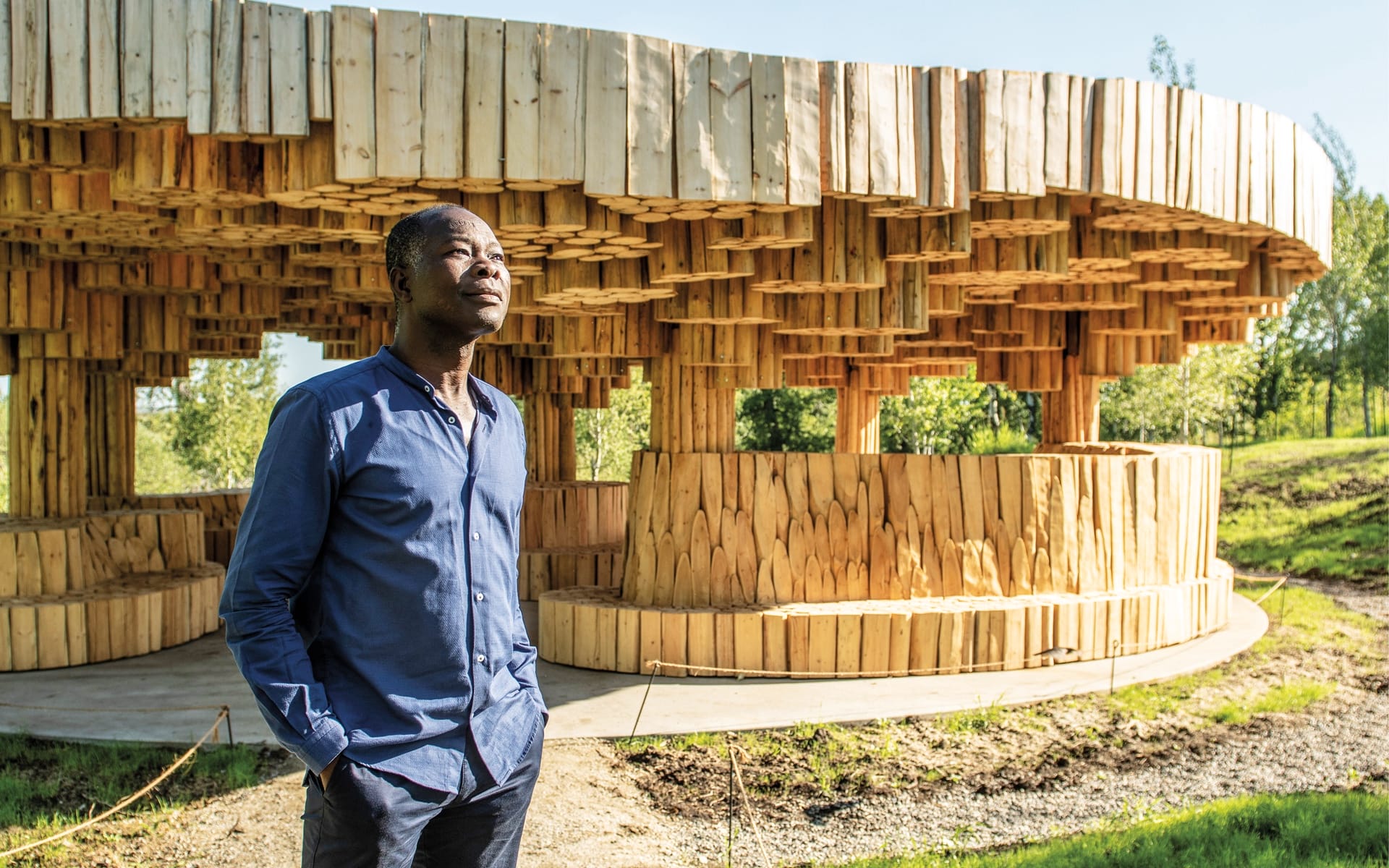 Person standing in front of a modern wooden art structure outdoors, with sunlight casting shadows on the ground.
