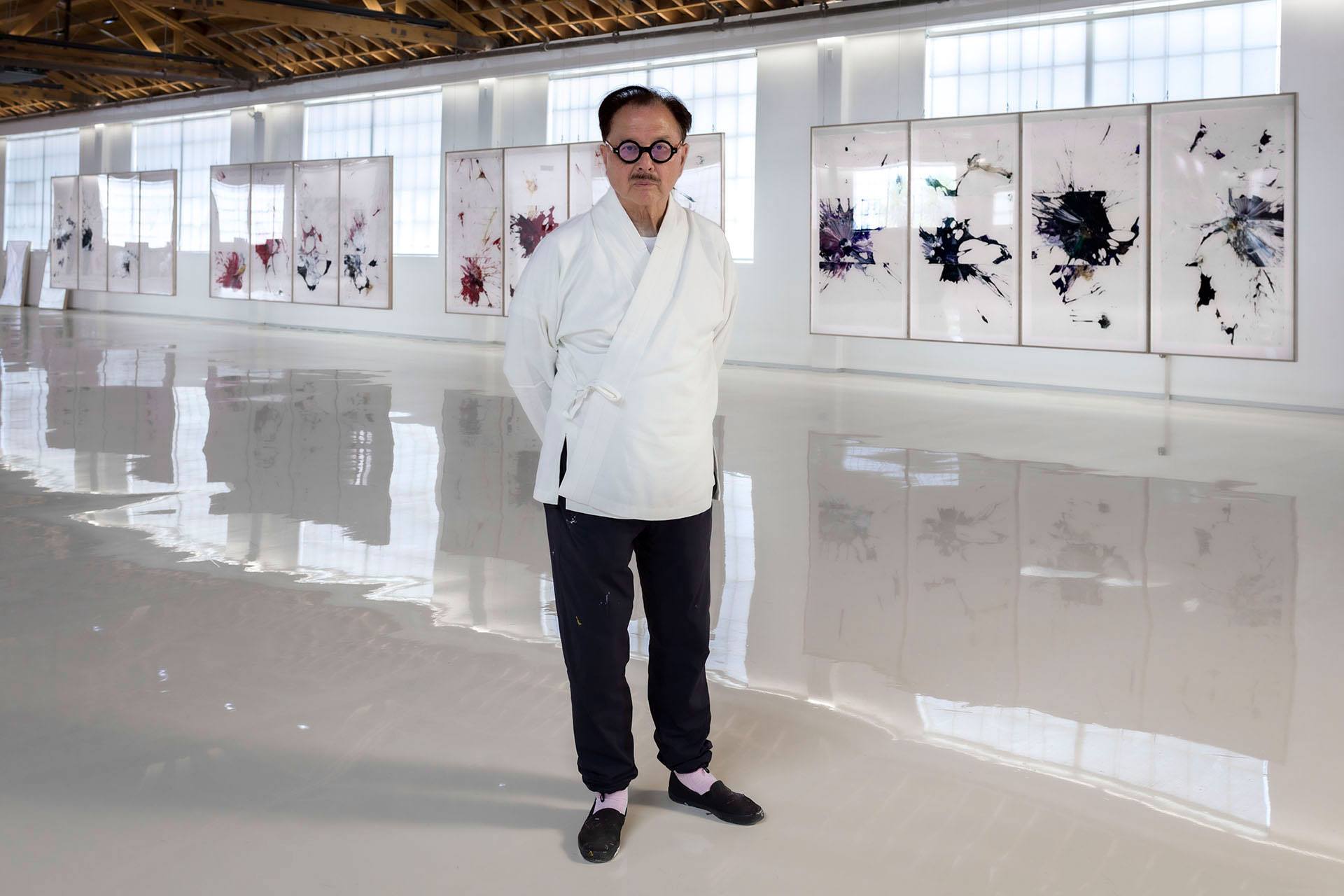 Artist standing in a modern gallery with abstract paintings displayed on the walls in the background.