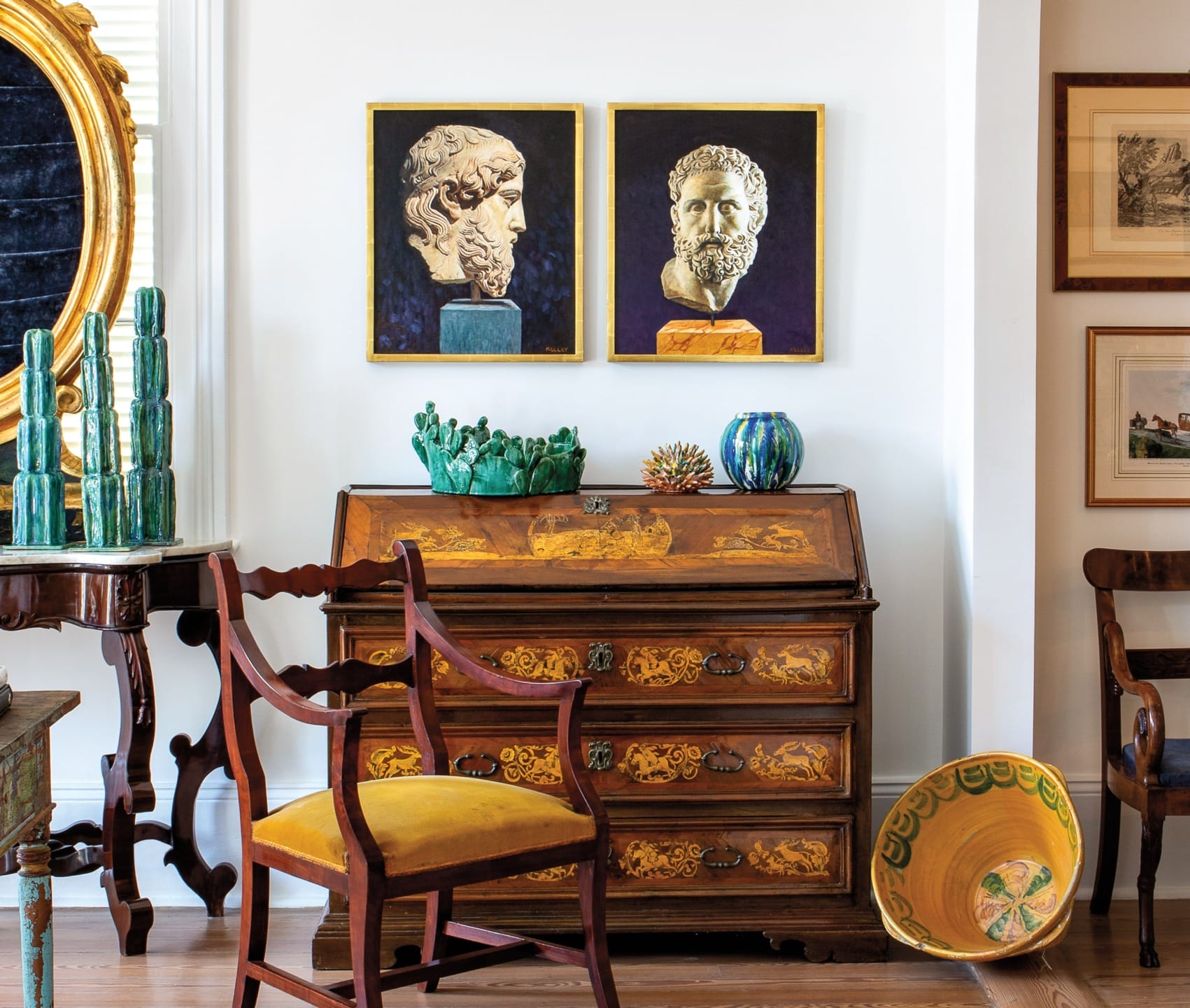 Vintage wooden desk with yellow upholstered chairs, colorful vases, classic paintings, and ornate mirror in eclectic room.