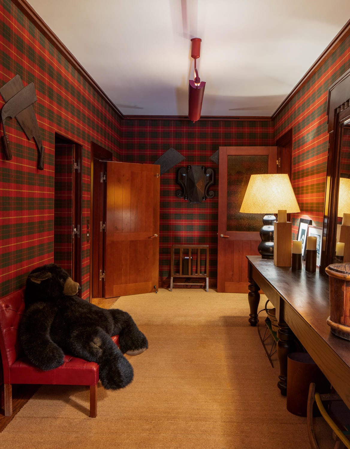 Room with red plaid wallpaper, wooden doors, a large teddy bear on a chair, a desk with a lamp, and decorative wall hangings.