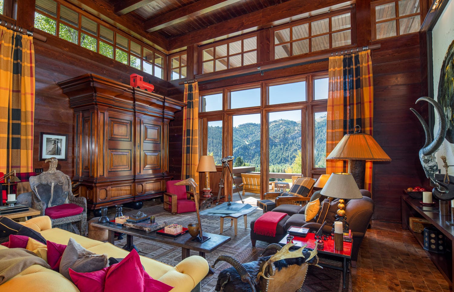 Cozy cabin living room with large windows, wooden furniture, colorful cushions, and a mountain view.
