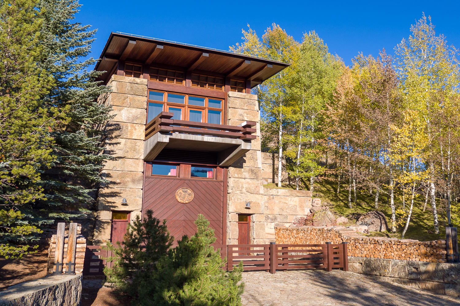 Two-story stone house with wooden accents surrounded by trees and blue sky in a tranquil setting.