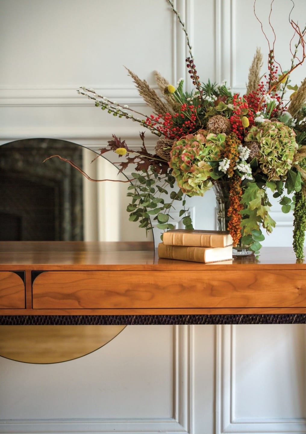 Decorative flower arrangement in a vase on a wooden table with books, set against a white paneled wall and mirror background.