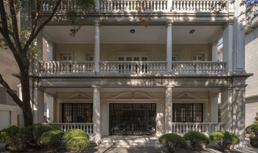 Exterior of a large, elegant residential building with columns, balconies, and well-maintained shrubs.