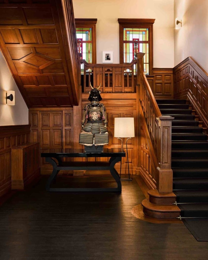 Samurai armor on display under wooden staircase with stained glass windows in a historical building interior.