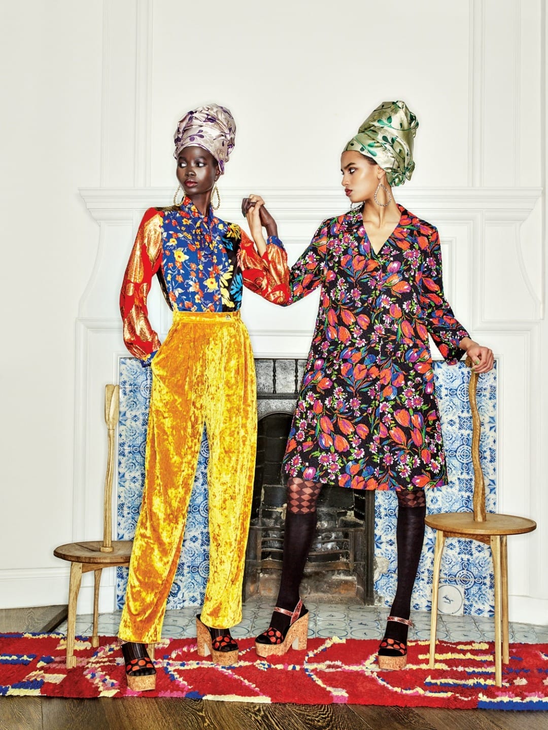 Two models in vibrant, patterned outfits and headwraps stand on a colorful rug in front of a decorative fireplace.