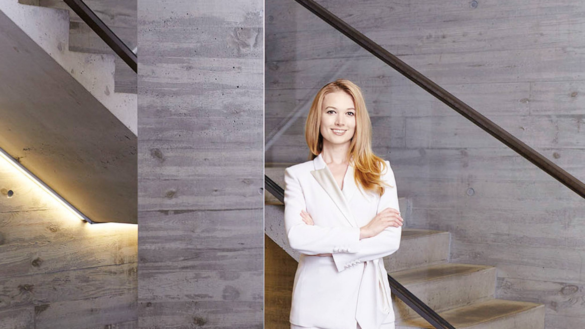 Woman in a white suit smiling, standing in a modern concrete stairwell with crossed arms.