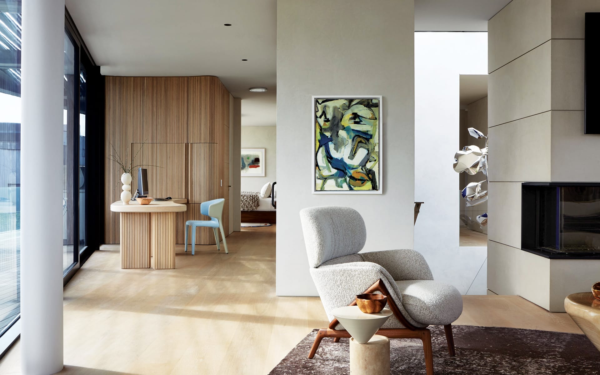 Modern living room with abstract art, stylish chair, and desk in open floor plan; natural light and wooden accents.