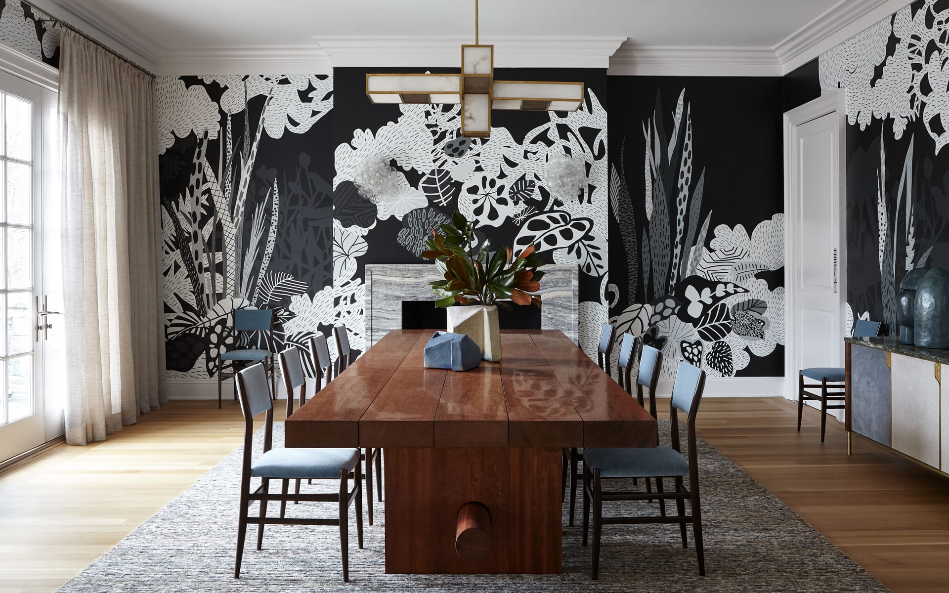 Dining room with a large wooden table, modern chandelier, and artistic black-and-white floral wall design.