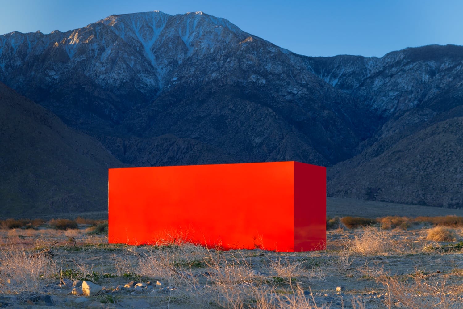 Red rectangular structure in a desert landscape with mountains in the background under a clear blue sky.