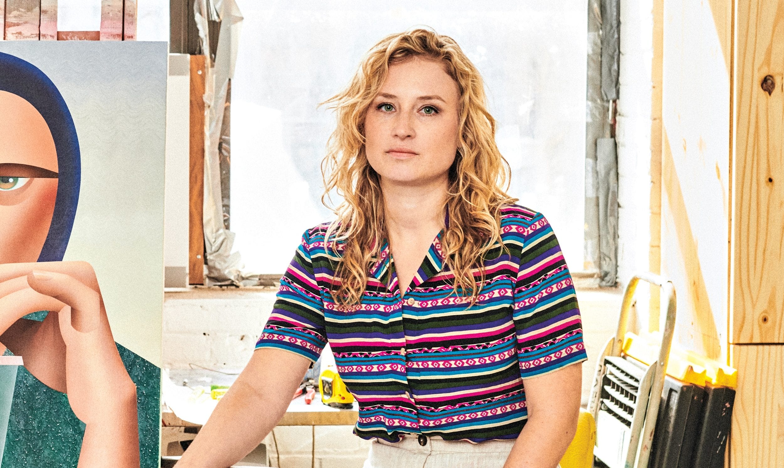 Person with curly hair in a colorful shirt standing by artwork in a bright studio.