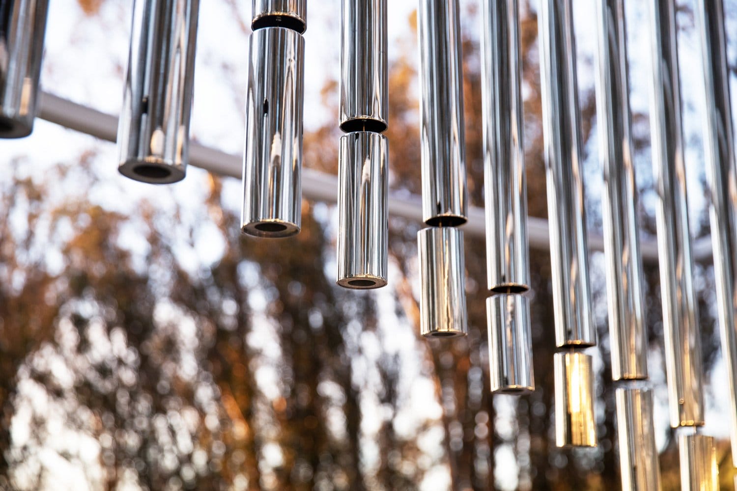 Metal wind chimes hanging outdoors with blurred trees in the background, reflecting sunlight at sunset.