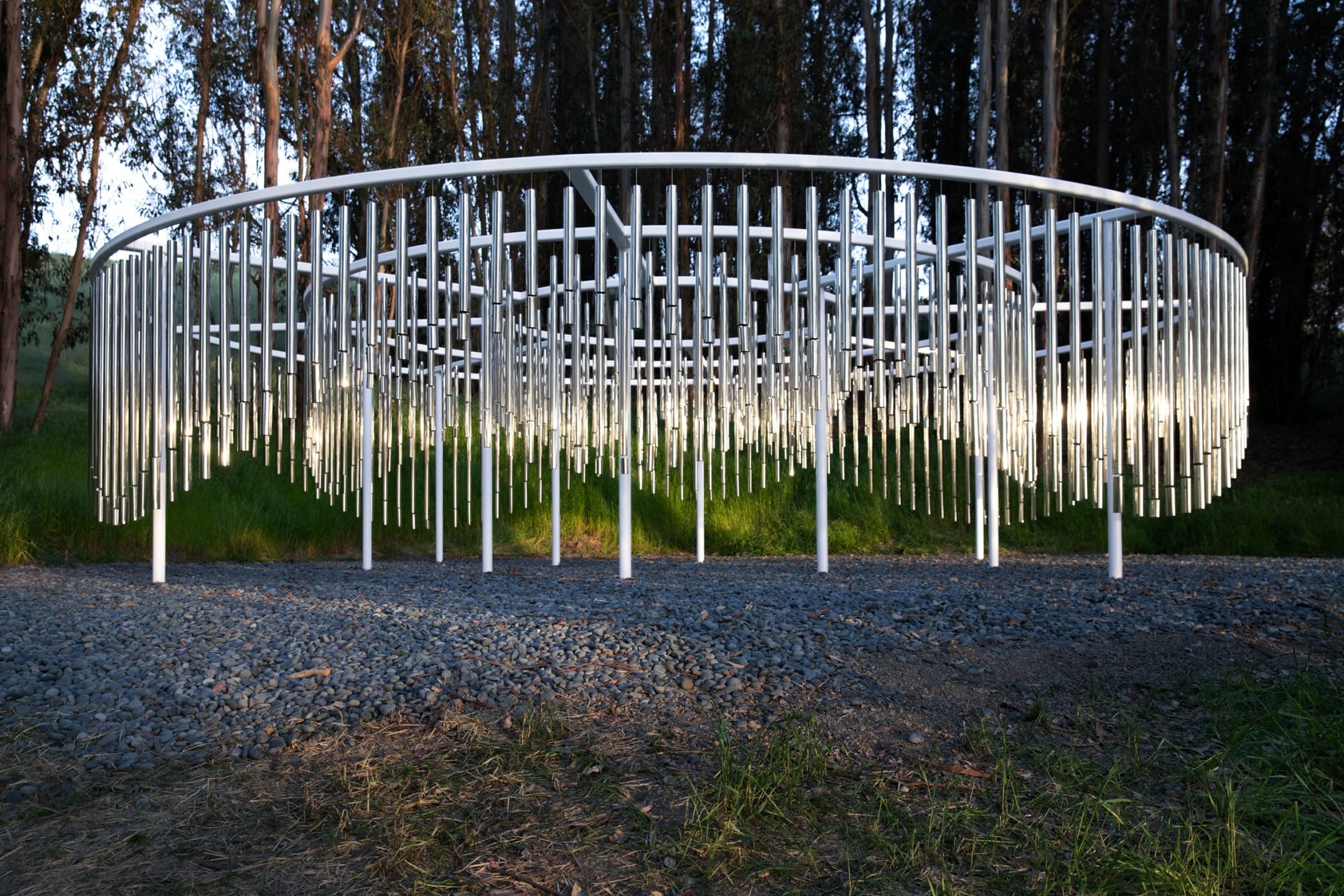 Circular metal sculpture with vertical rods, set in a forested area with sunlight filtering through the trees.