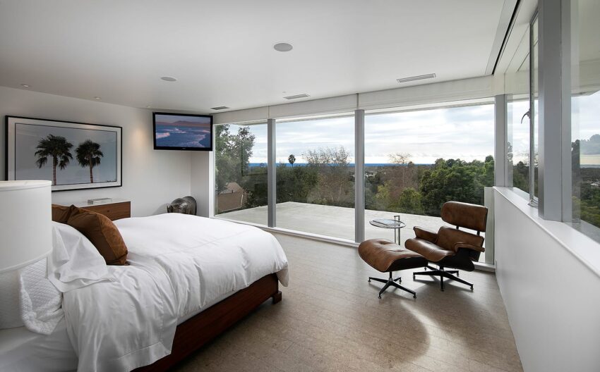Modern bedroom with large windows, white bedding, and a cozy lounge chair overlooking a scenic outdoor view.