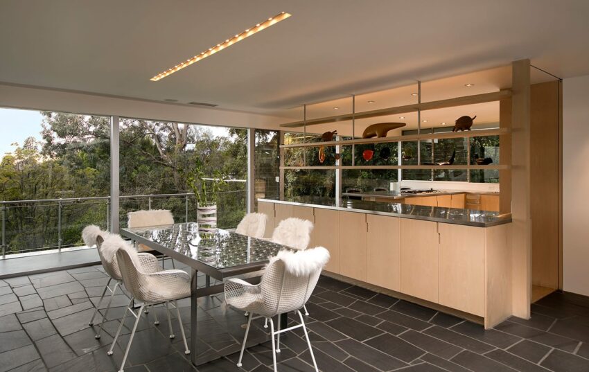 Modern dining area with glass table, white chairs, large windows, and view of lush greenery outside.