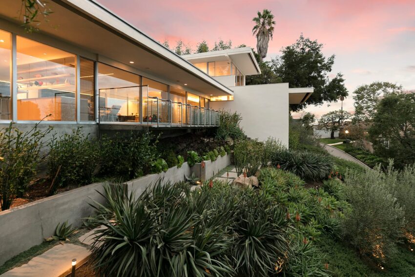 Modern house with large glass windows and lush landscaping at sunset.