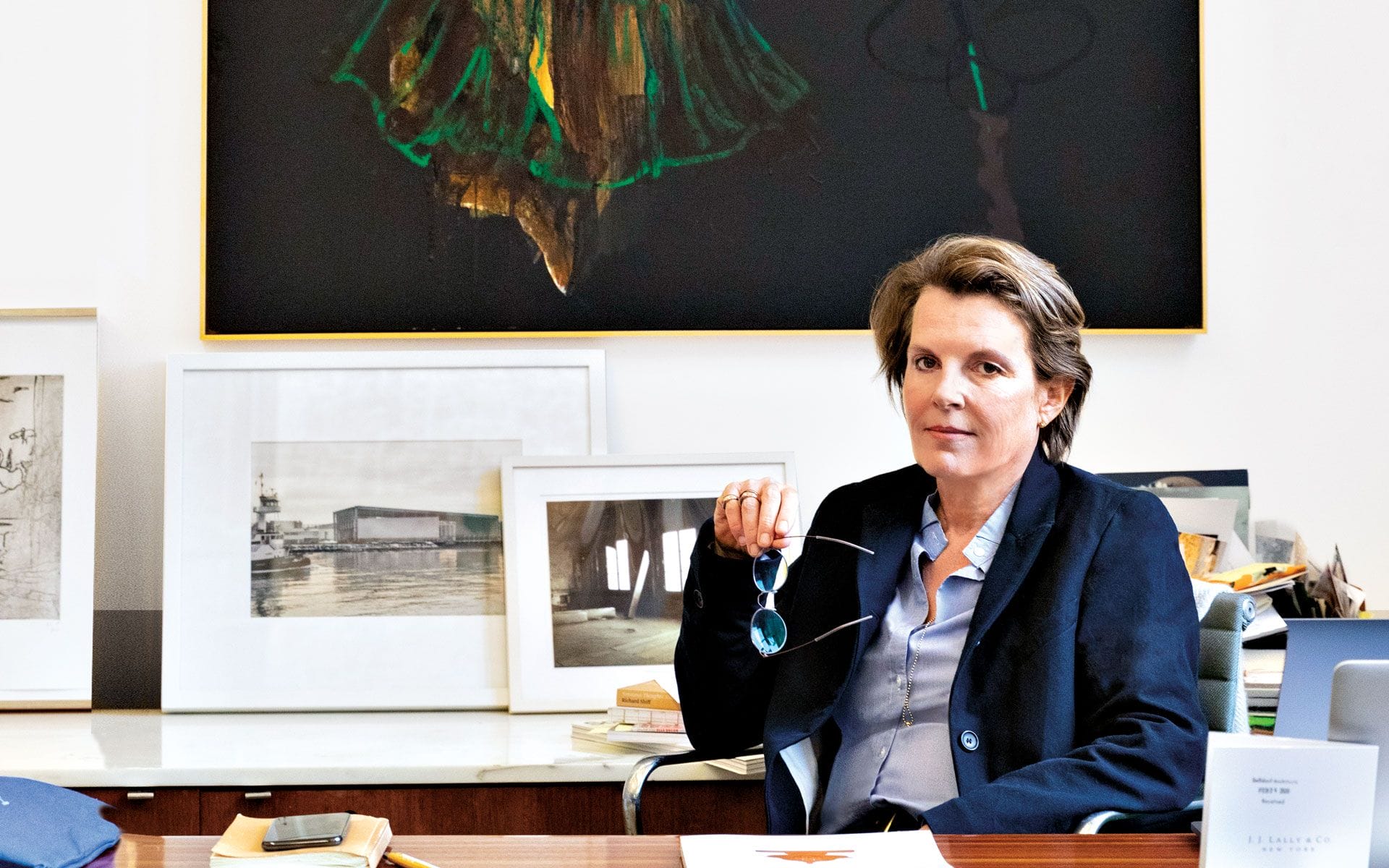 Person sitting at a desk in an art-filled room, holding glasses and wearing a blazer, with framed artworks on the wall.