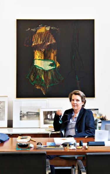 Person sitting at a desk with art on the wall behind, holding an object. Office setting with various items on the desk.