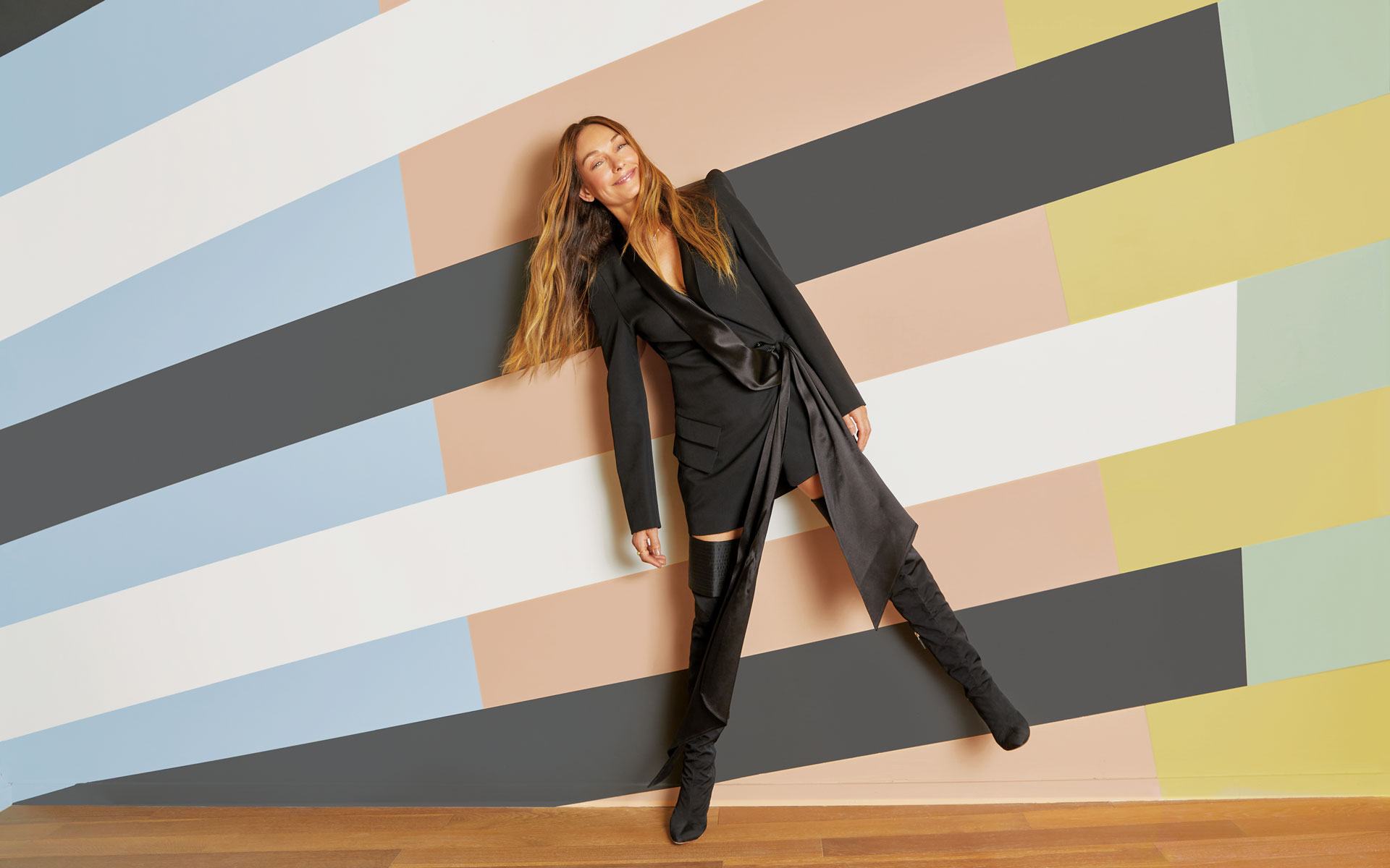 Person leaning on a colorful, striped wall wearing a black outfit and knee-high boots.