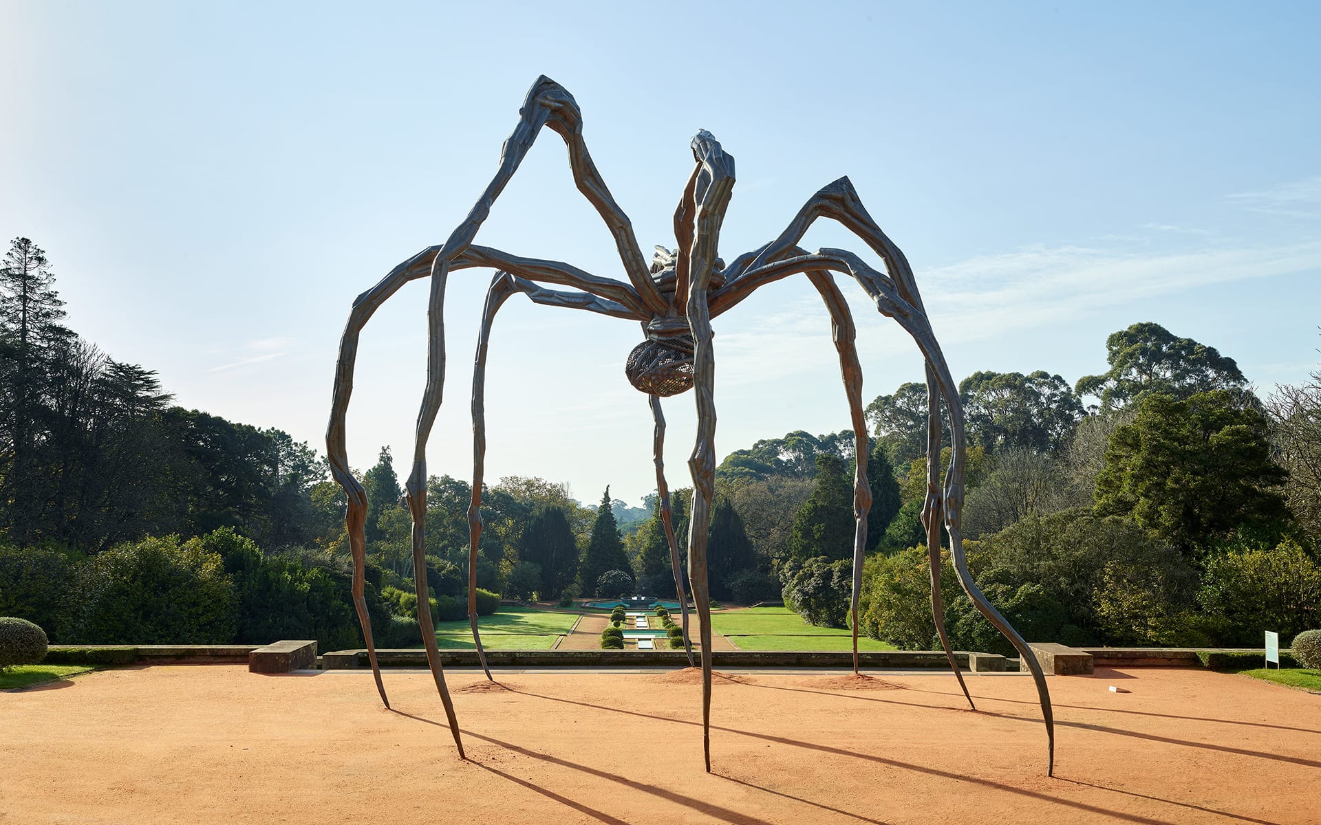 Large metal spider sculpture in a garden setting with trees and a clear sky in the background.