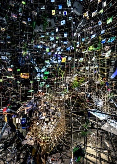 Art installation with numerous suspended electronic devices and images creating a complex, colorful network-like structure.