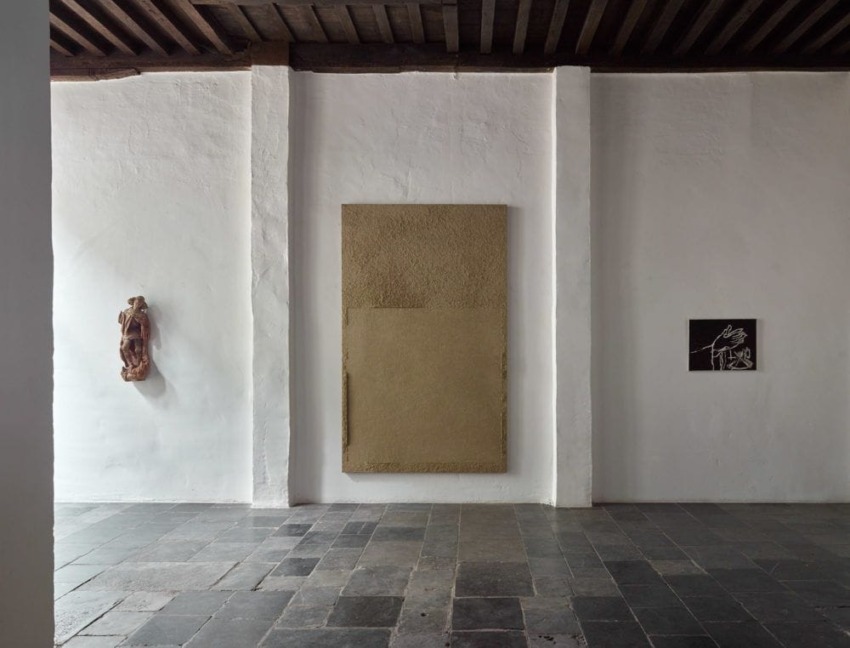 Minimalist art display in a gallery with textured beige canvas, small sculpture, and wall-mounted artwork on white walls.