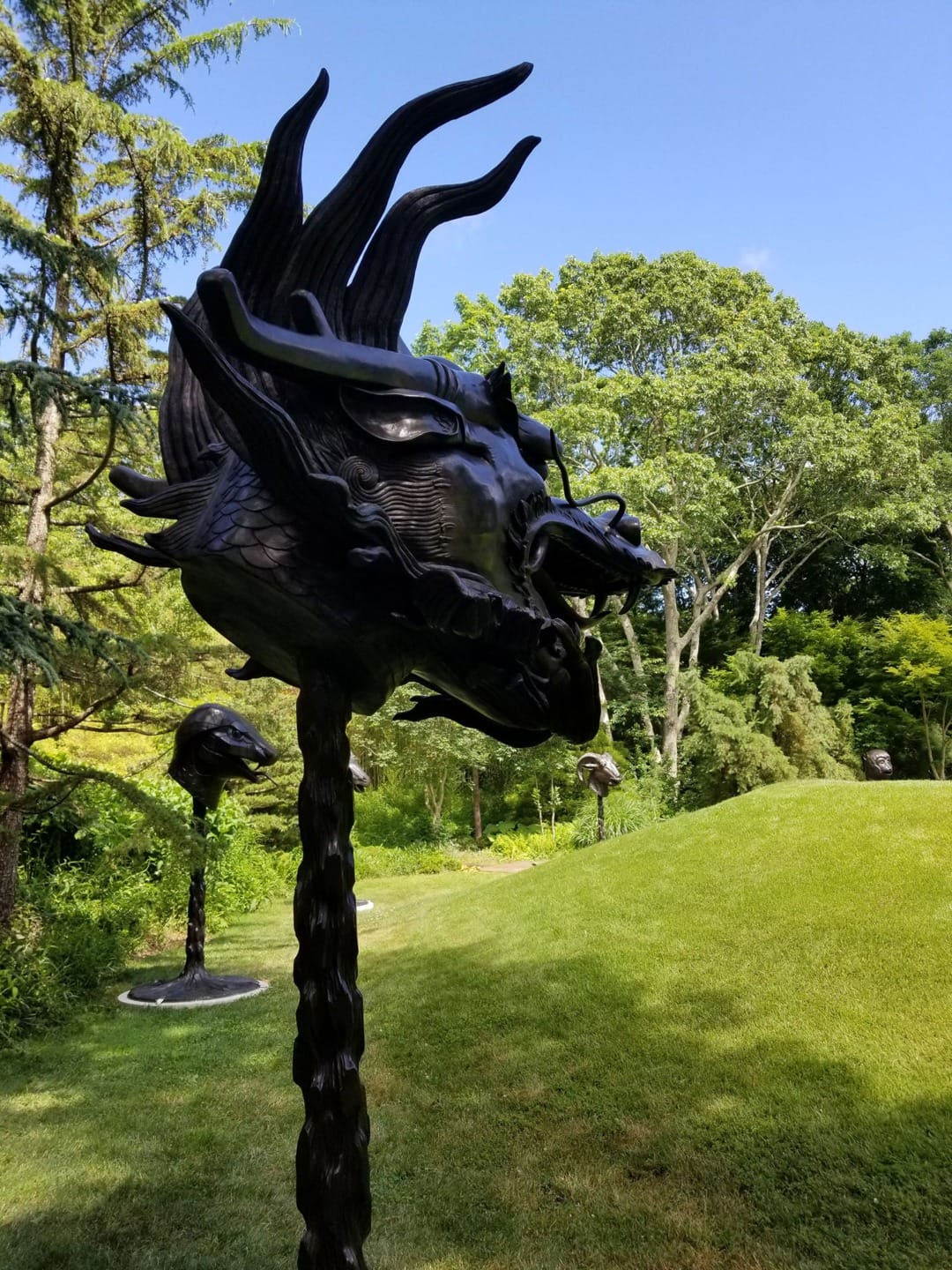 Sculpture of a dragon head with flowing mane in a lush green outdoor setting under a clear blue sky.