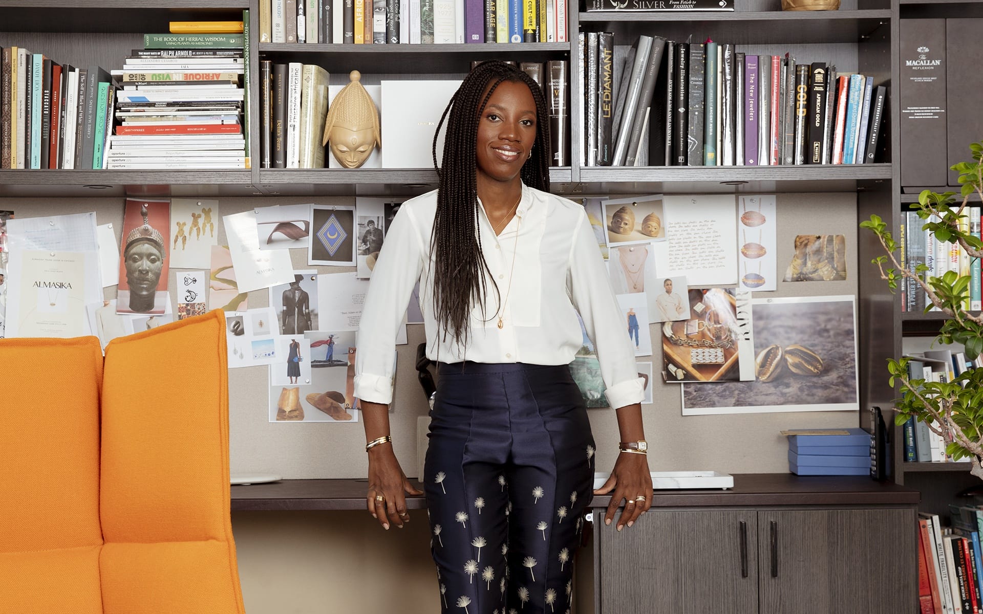 Person standing in an office with bookshelves and an orange chair, wearing a white shirt and patterned pants.