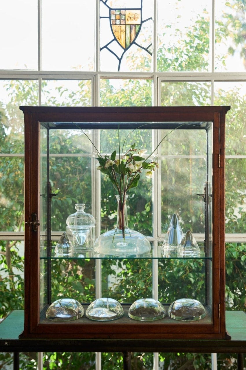 Vintage glass cabinet with a vase of flowers and various glass objects, set against a window with a stained glass accent.