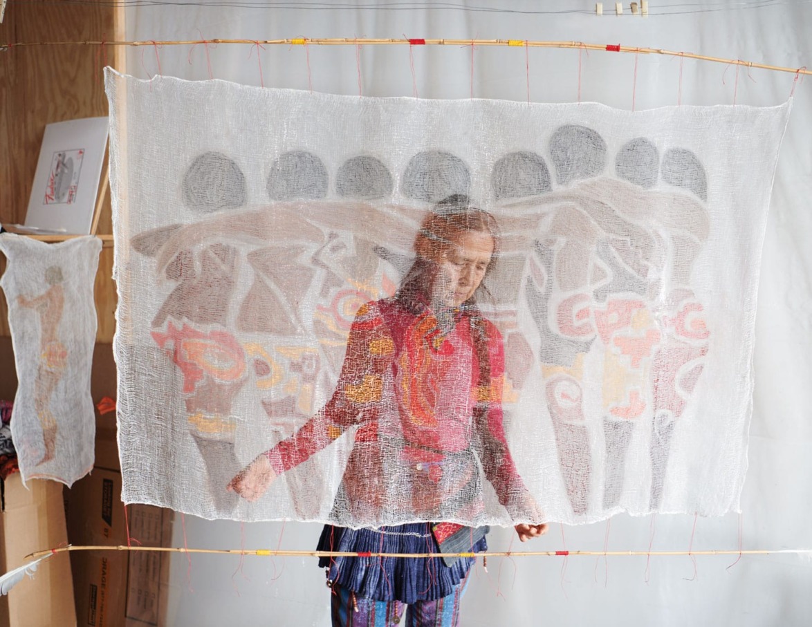 Woman standing behind a translucent fabric with abstract art in the background, creating an artistic layered effect.