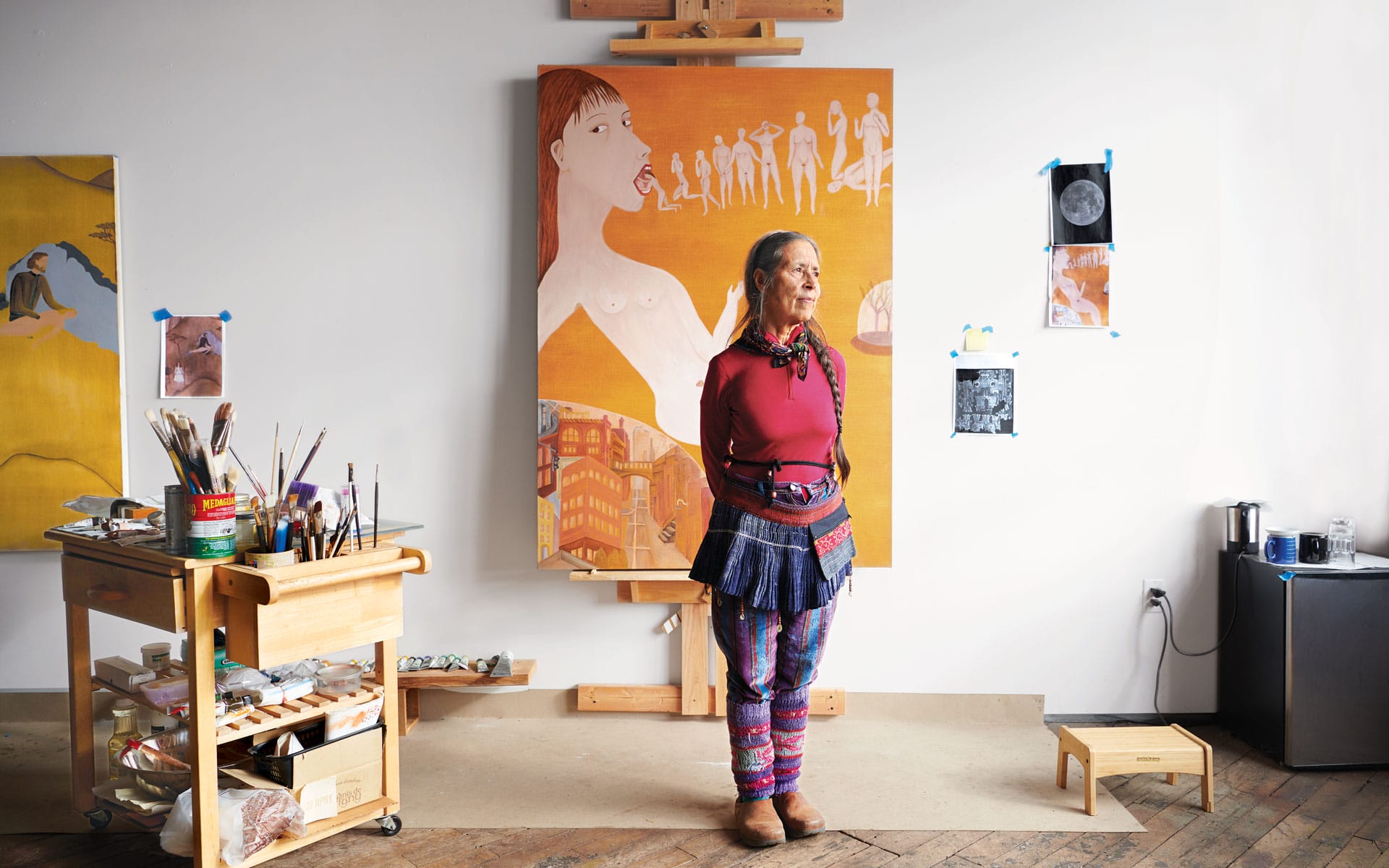 Artist standing in studio with colorful attire, surrounded by art supplies and canvas paintings on easels.