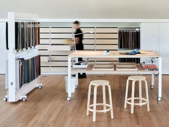 Modern workspace with fabric samples on racks, a long table, and two stools on a wooden floor.