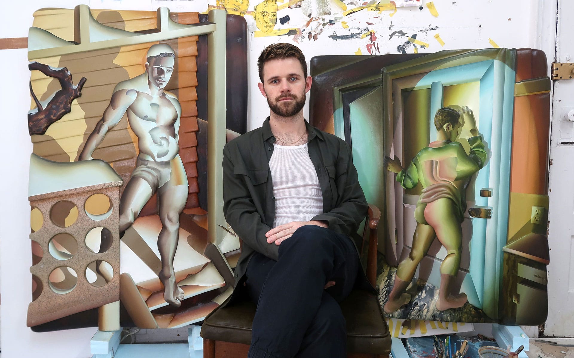 Artist seated between two colorful, abstract paintings featuring human figures, set in a well-lit studio space.