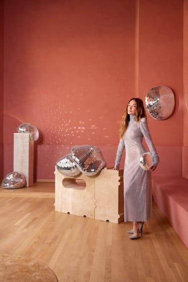 Woman in a sparkling dress stands near a table with reflective sculptures in a modern room with red walls.
