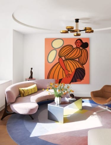 Modern living room with abstract art, curved sofa, geometric rug, colorful flowers in vase, and contemporary lighting fixture.