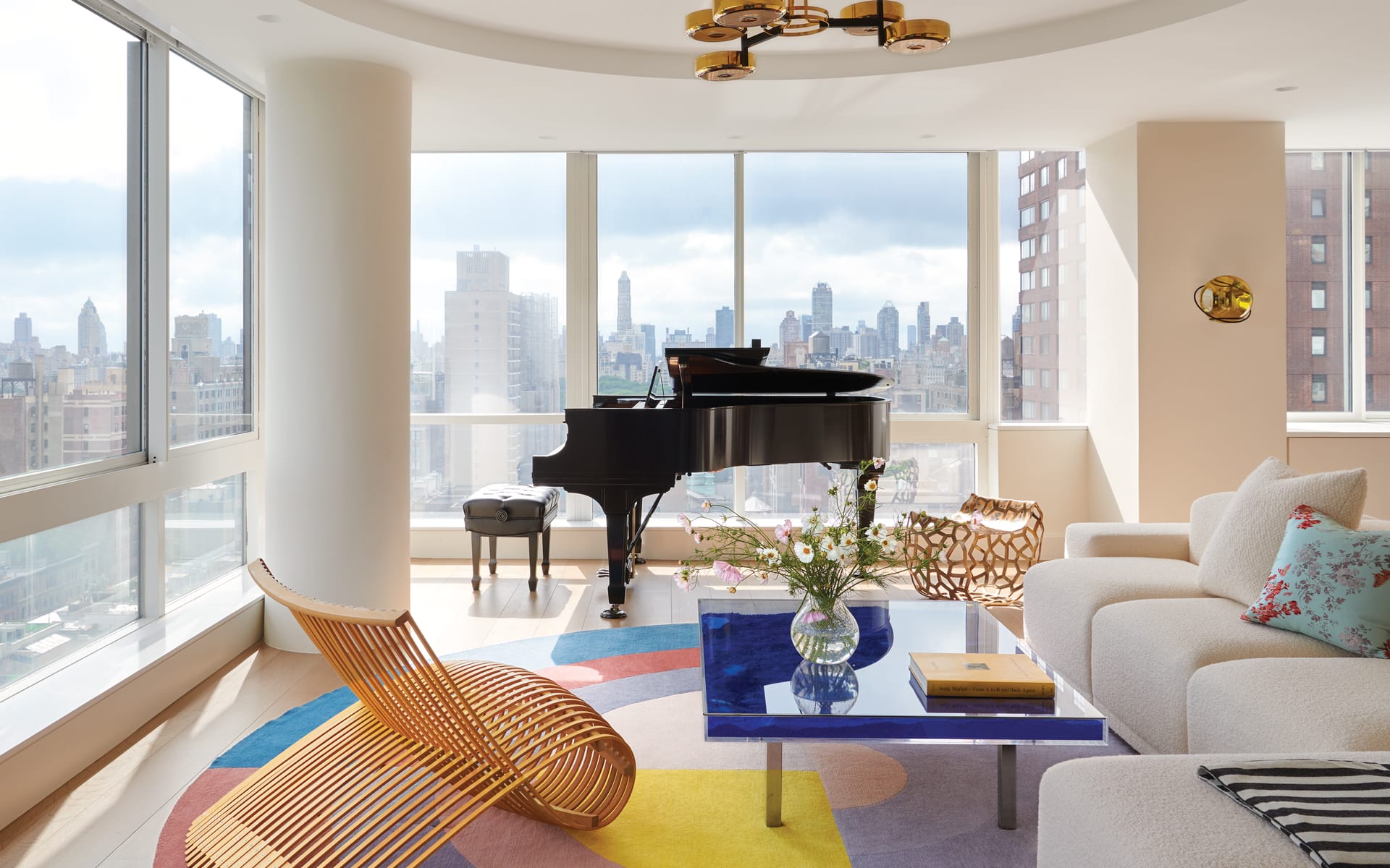 Modern living room with large windows, grand piano, unique furniture, colorful rug, and city skyline view.