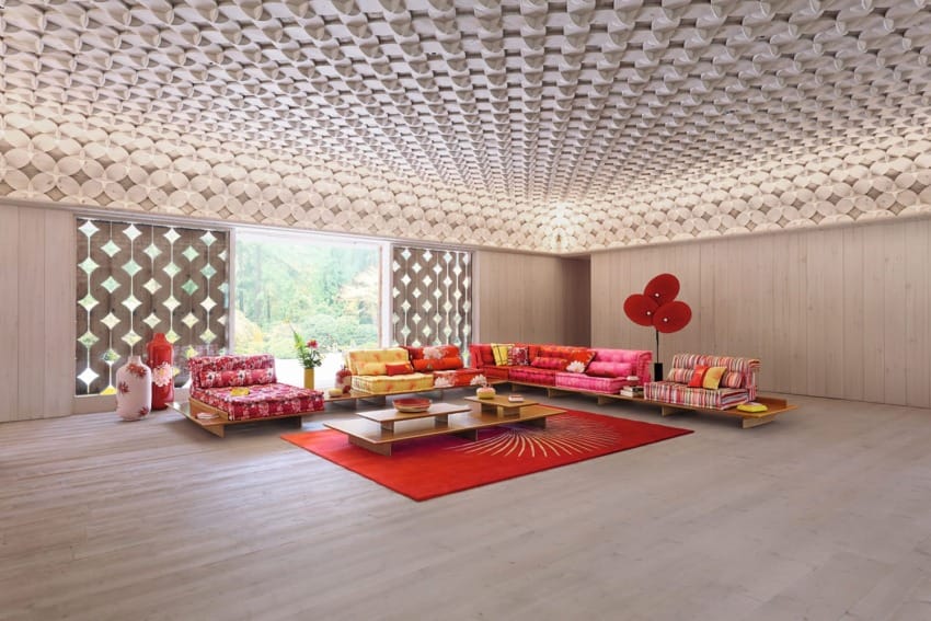 Modern living room with colorful sofas, red rug, geometric ceiling, and large windows showcasing a garden view.