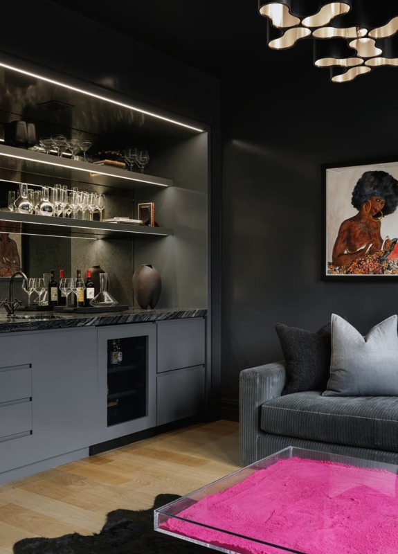 Modern living room with dark walls, a bar area with glassware, a sofa with cushions, and a pink rug on a wooden floor.