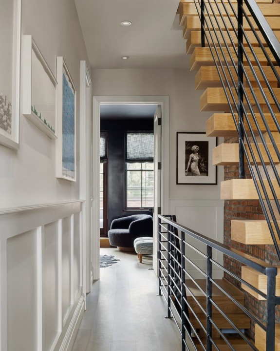 Modern hallway with wooden stairs, black railing, and artwork on walls leading to a cozy room with a chair by the window.