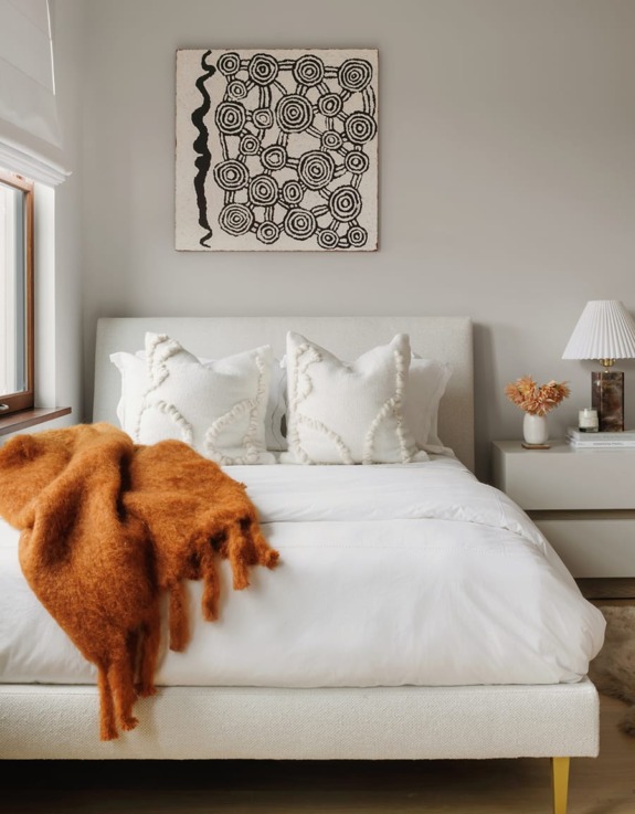 Cozy bedroom with white bed, two pillows, orange throw blanket, bedside table, lamp, and abstract wall art.