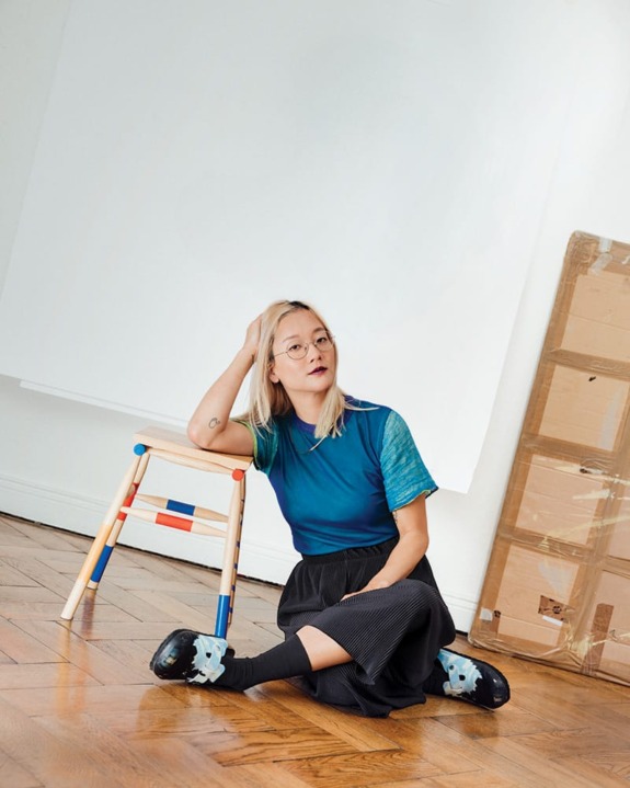 Person sitting on the floor beside a colorful stool in a well-lit room with wooden flooring and packaging materials in the background.