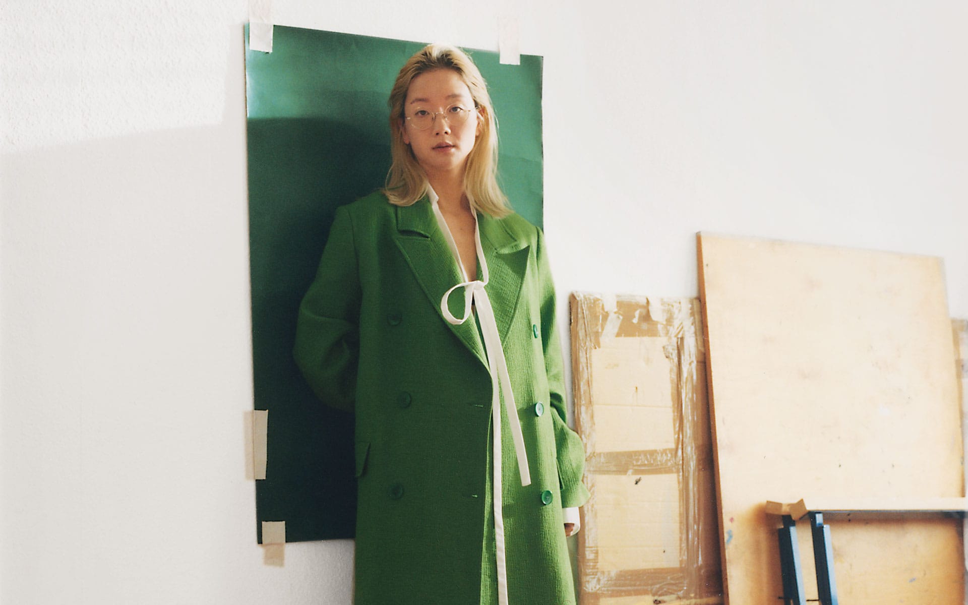 Person wearing a green coat stands indoors against a large green paper background in an artist's studio.