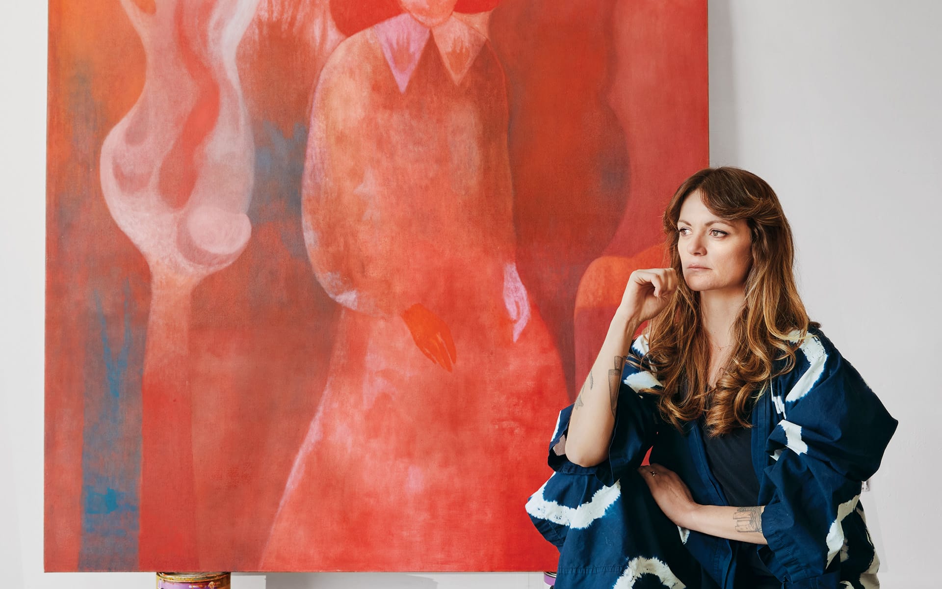 Artist sitting pensively in front of a large red painting with abstract human figures in a studio setting.