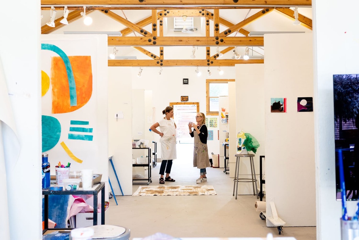 Artists discussing in a bright, modern studio with colorful paintings and wooden beams.