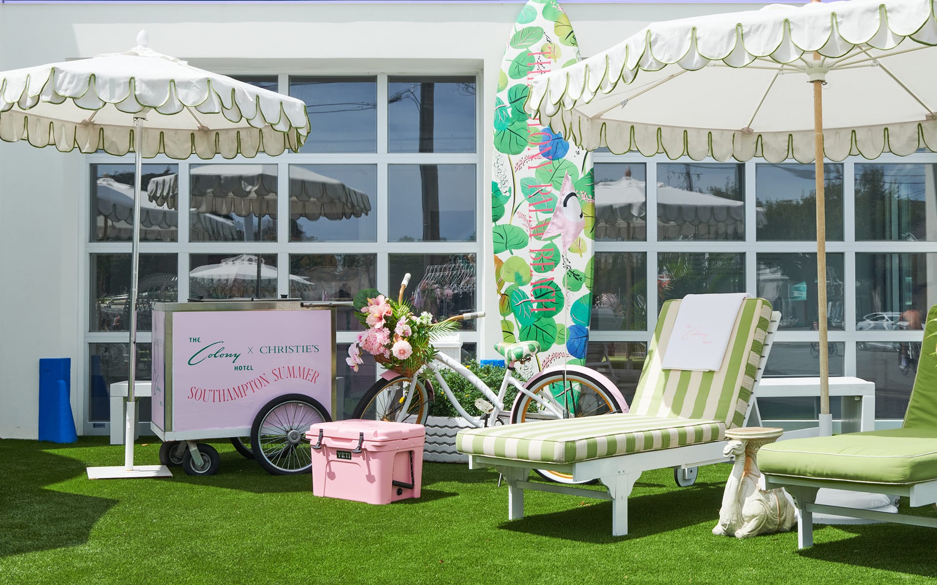 Outdoor seating area with pink bike, surfboard, and flowers under white umbrellas on a sunny day.