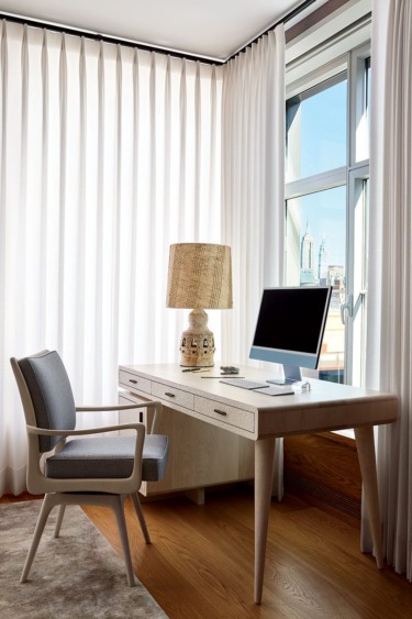 Minimalist home office with wooden desk, chair, computer, and lamp by a large window with white curtains.