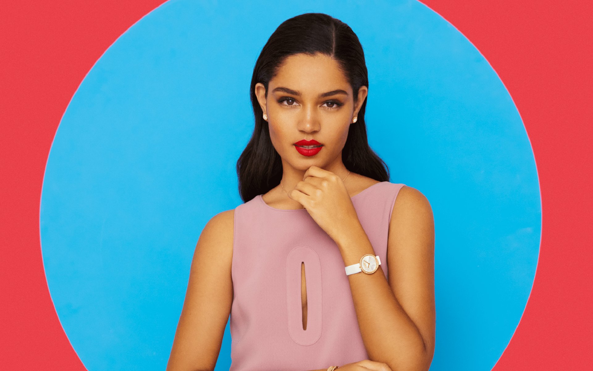 Woman in pink sleeveless dress with a watch, posing in front of a blue and red background